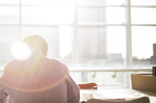view of a businessman from behind, sitting at a desk with sun shining through large windows