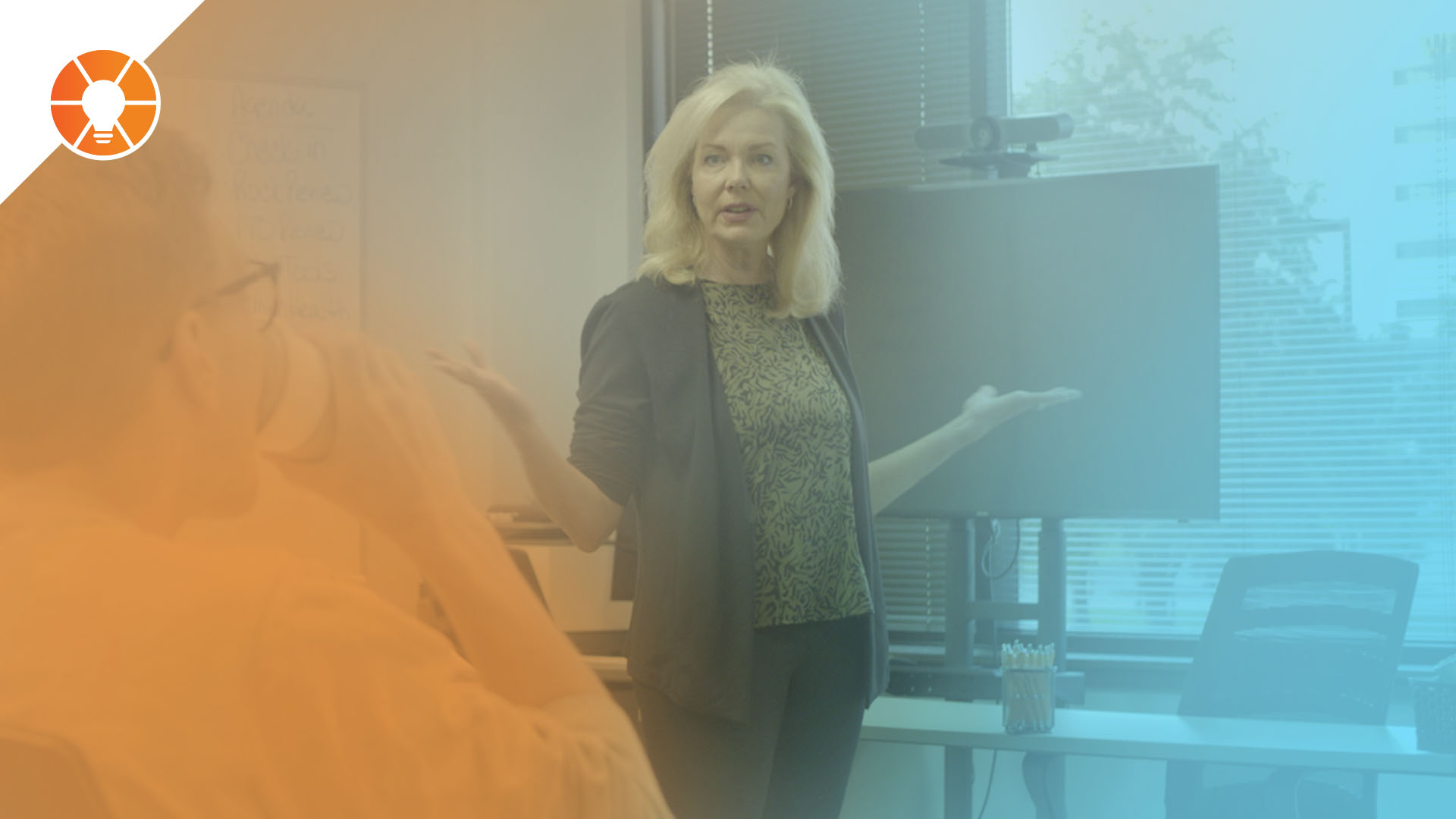 A business leader stands at the front of a conference room, gesturing as she speaks to colleagues during a meeting, with a blurred attendee in the foreground and a presentation screen behind her, overlaid with a blue-to-orange gradient.