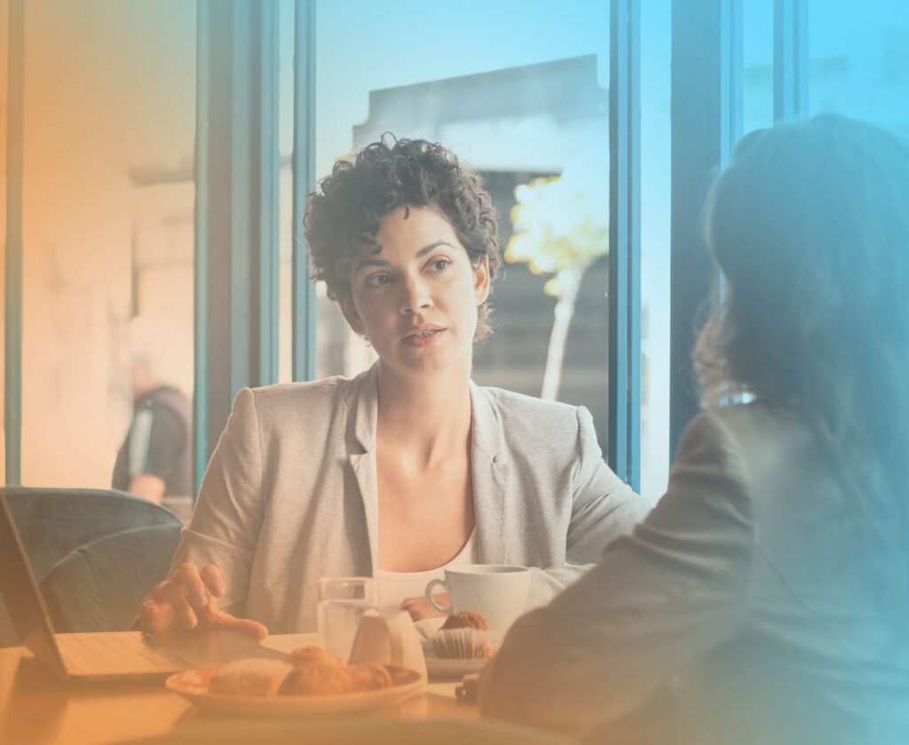Two business professionals sit across from each other at a café-style table, having a focused one-on-one conversation beside a laptop, coffee cups, water glasses, and pastries, with a warm orange-and-blue overlay across the image.