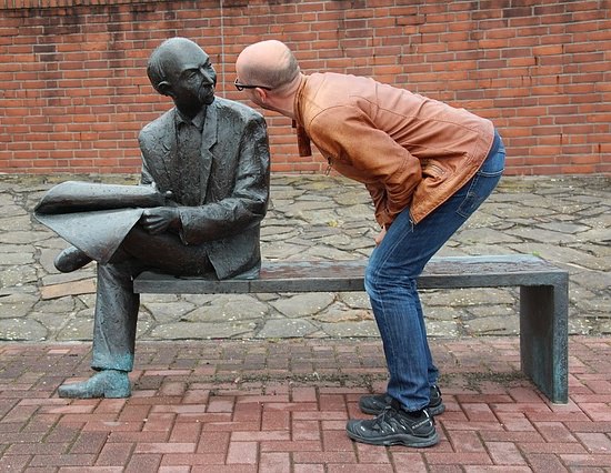 man standing face-to-face with a statue of a man outside on a bench
